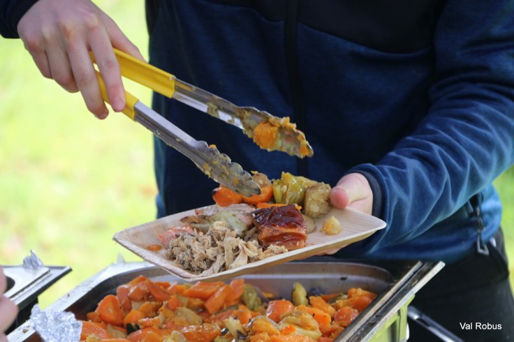 A person using tongs to serve food on a wooden plate, featuring roasted vegetables and meat from a large serving tray.