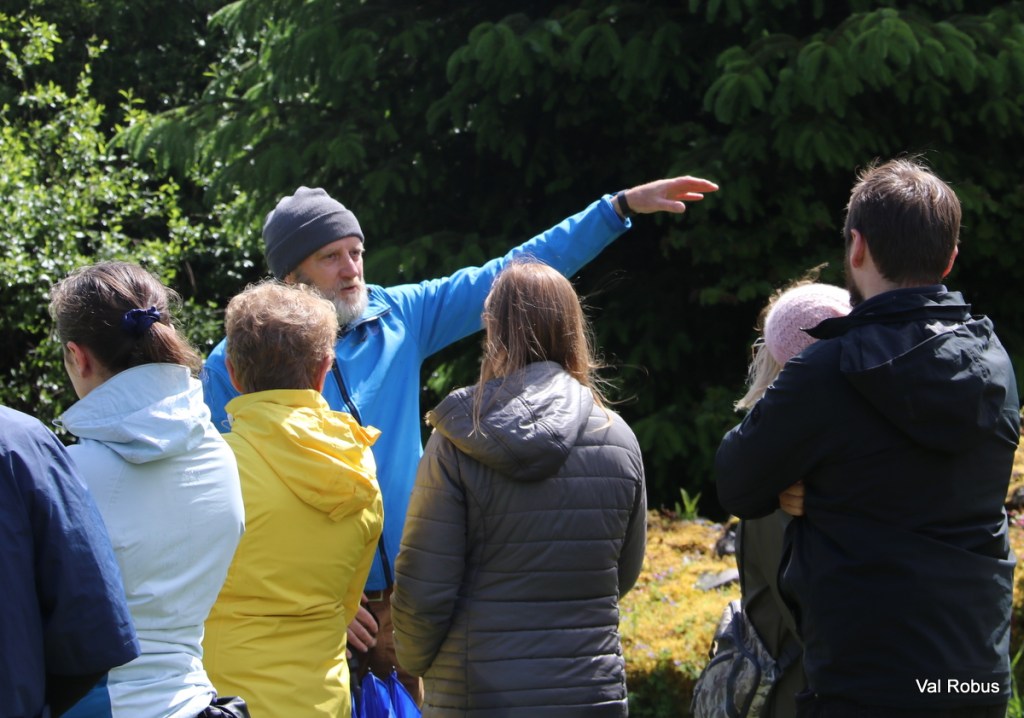 A guide explaining something to a group of people in a natural setting, with greenery in the background.