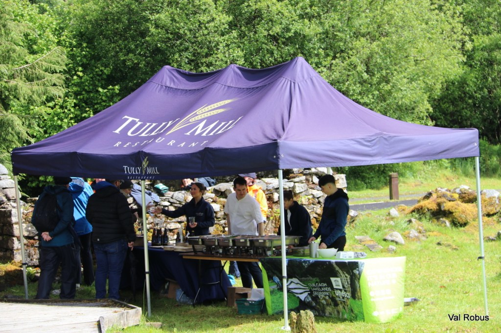 A pop-up tent with the logo of Tully Mill Restaurant, set in a green outdoor area with people gathered around a table under the tent.