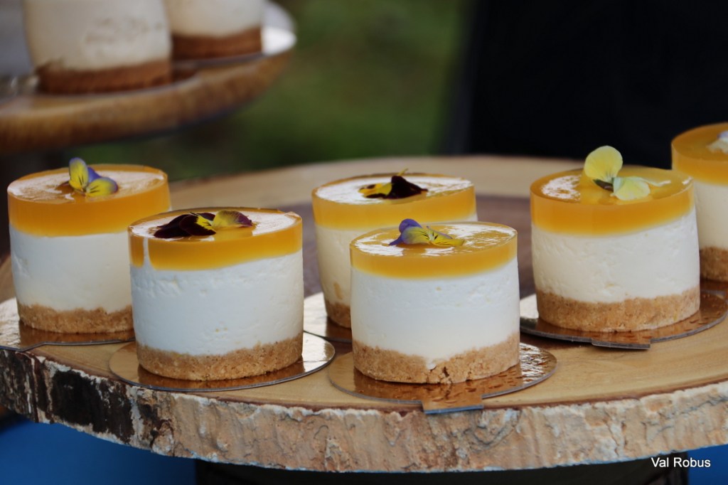 Several individual cheesecakes with a yellow fruit topping and edible flower decorations placed on a wooden platter.