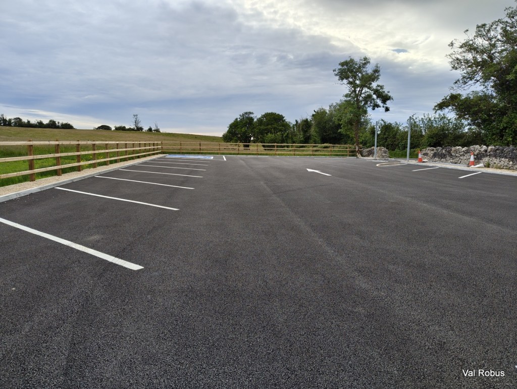 An empty parking lot with marked spaces, surrounded by a wooden fence and greenery, under a cloudy sky.