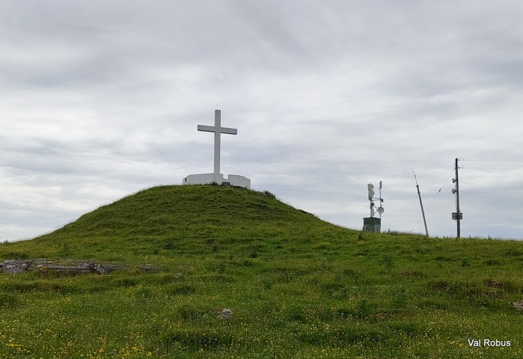 A large white cross on a grassy hill, with telecommunications equipment in the background and a cloudy sky.