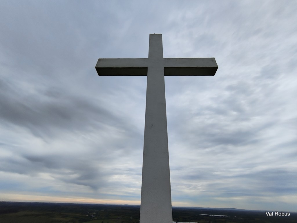 A large white cross against a cloudy sky, with a view of the landscape in the background.
