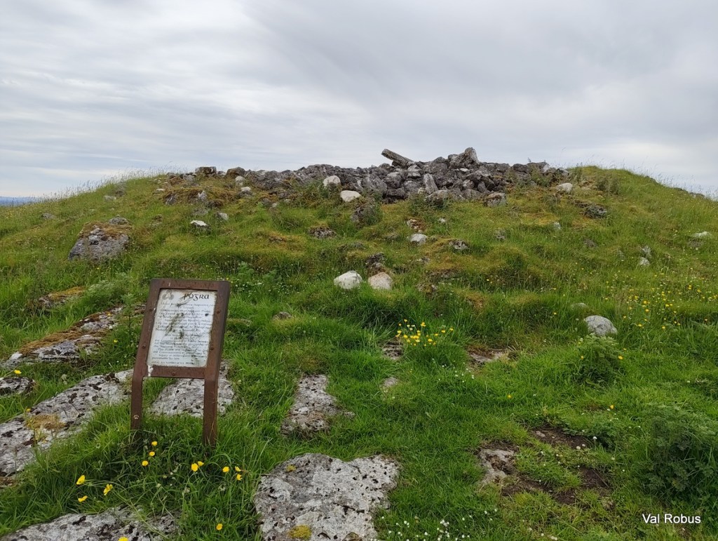 A grassy hilltop featuring a stone structure and a weathered information sign, surrounded by wildflowers and cloudy skies.