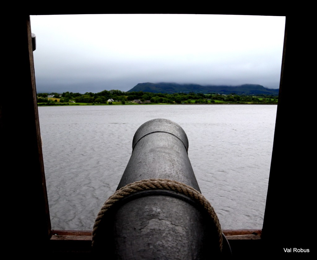 View through a cannon pointing out over a river towards a mountainous landscape under a cloudy sky.