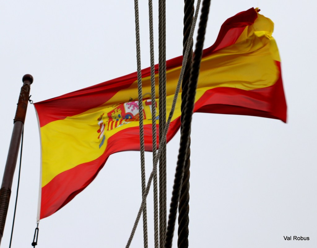 Spanish flag waving against a gray sky, with ropes in the foreground.