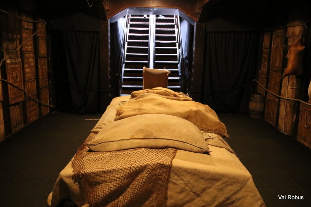 Interior view of a dimly lit space resembling a ship's hold, featuring a bed with burlap sacks and pillows, and stairs leading up to a brighter area.