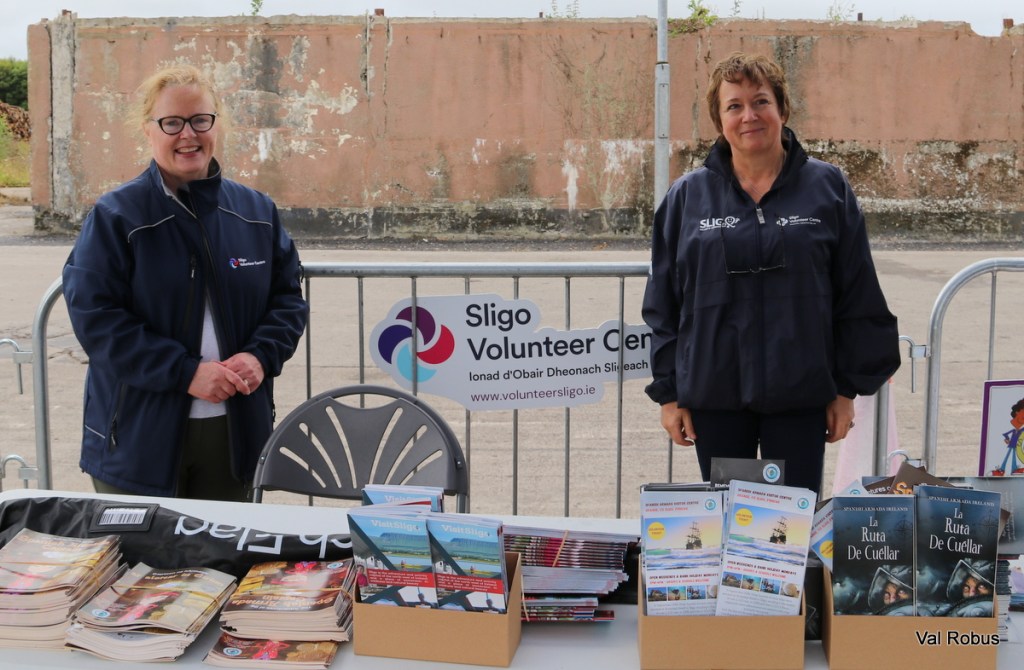 Two women standing behind a table at an outdoor event, promoting the Sligo Volunteer Center. The table is adorned with brochures and promotional materials, and a banner for the center is displayed in the background.