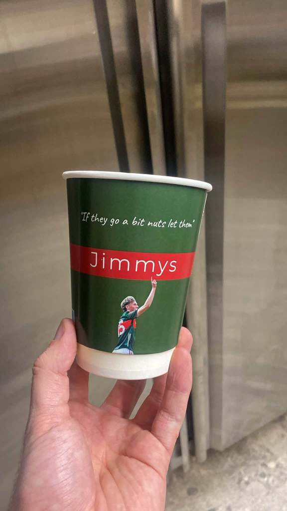 A hand holding a green cup with a red band reading 'Jimmys' and a quote that says, 'If they go a bit nuts let them.' There is an illustration of a person in a sports uniform on the cup.