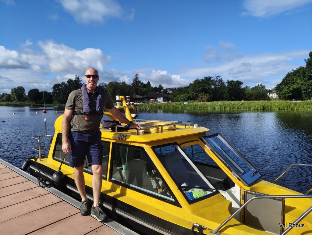 A man in sunglasses and a life jacket stands on the deck of a yellow boat by a calm lake, surrounded by greenery and a blue sky with clouds.