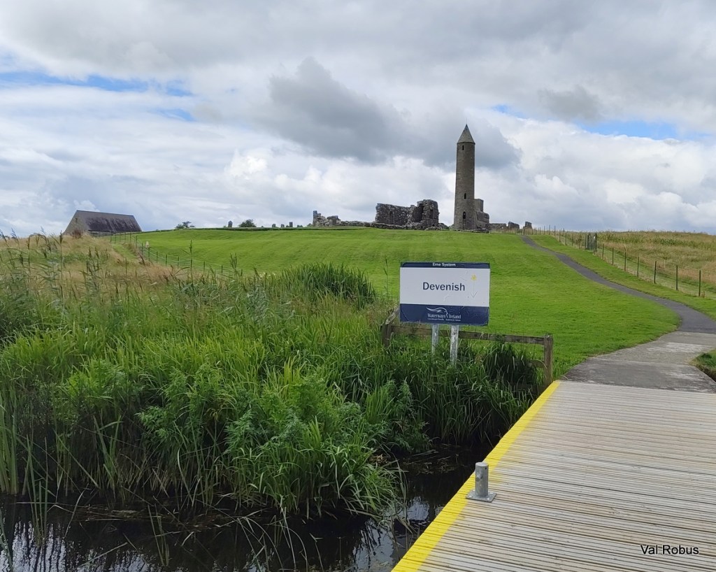 Scenic view of Devenish Island, featuring a signpost, a round tower, and ruins in the background, surrounded by greenery and a pathway.