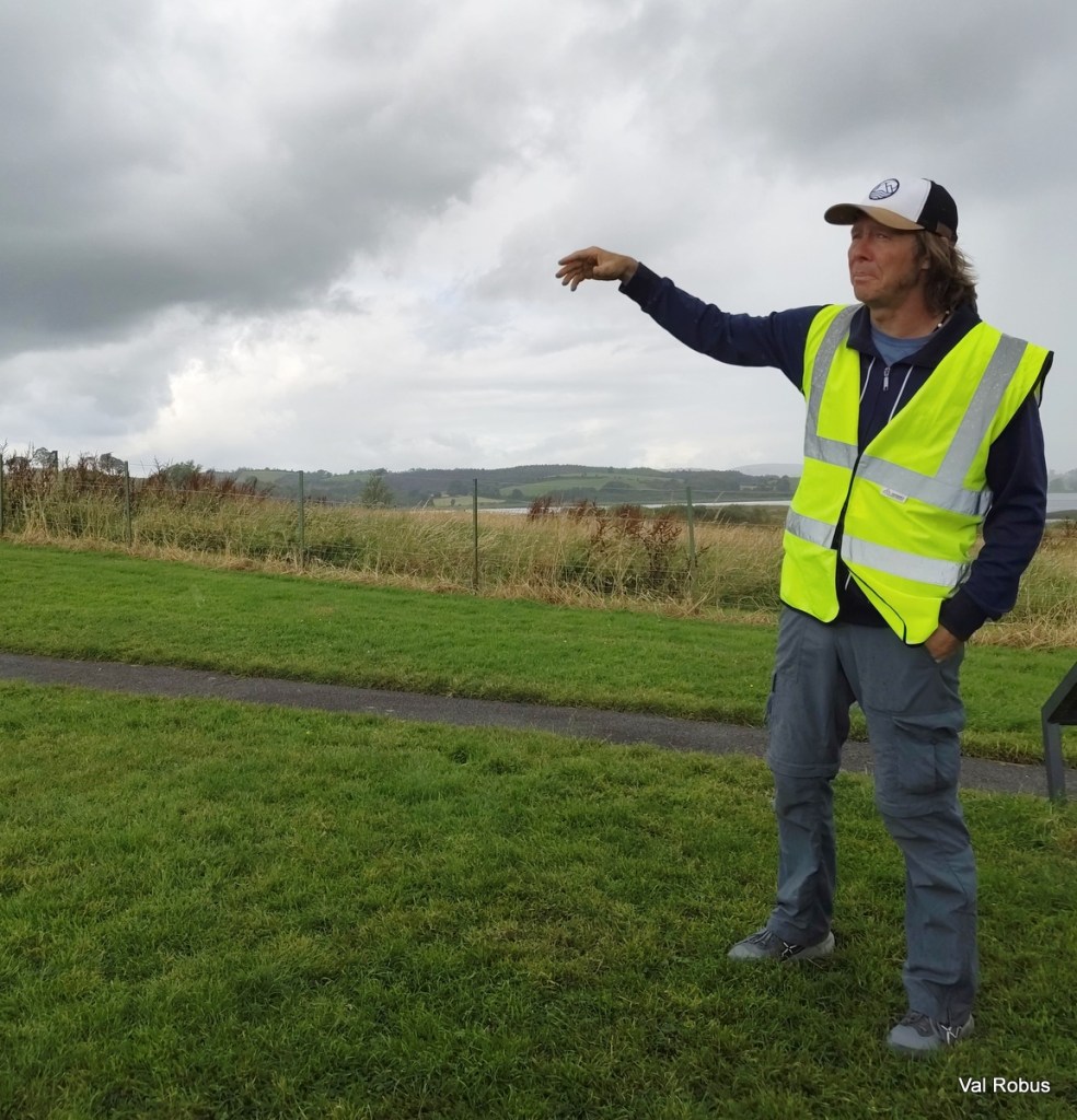 A man wearing a high-visibility vest and a cap stands outdoors, gesturing with his right hand towards a cloudy sky and a landscape of grass and hills in the background.