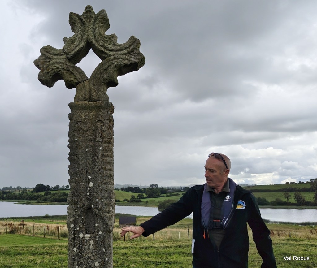 A man stands next to a tall, intricately carved stone cross with leafy designs, set against a backdrop of cloudy skies and a green landscape near a body of water.