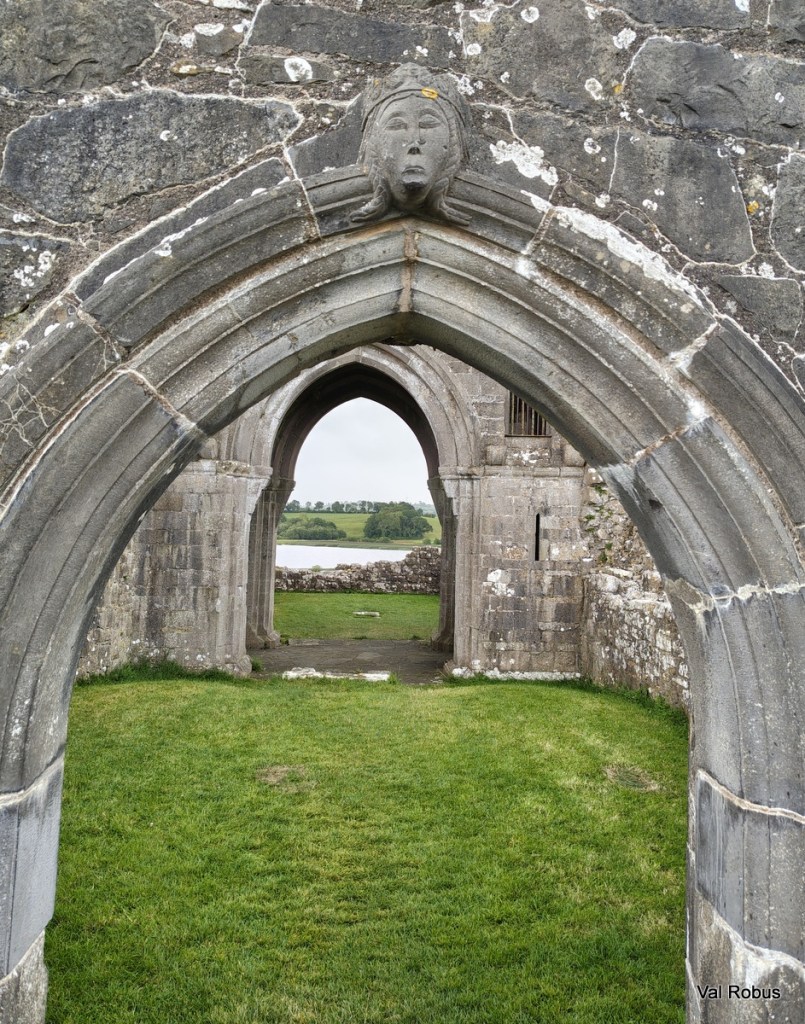 A stone archway with a carved face at the top, leading to a grassy area and an open view of fields beyond.