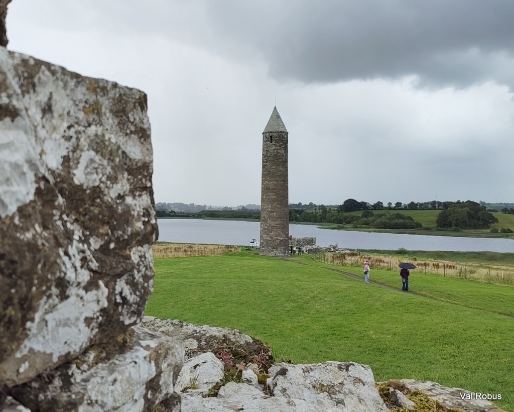 A stone tower stands near a lake, with grassy fields and a cloudy sky in the background. Two people are walking along a path, one holding an umbrella.