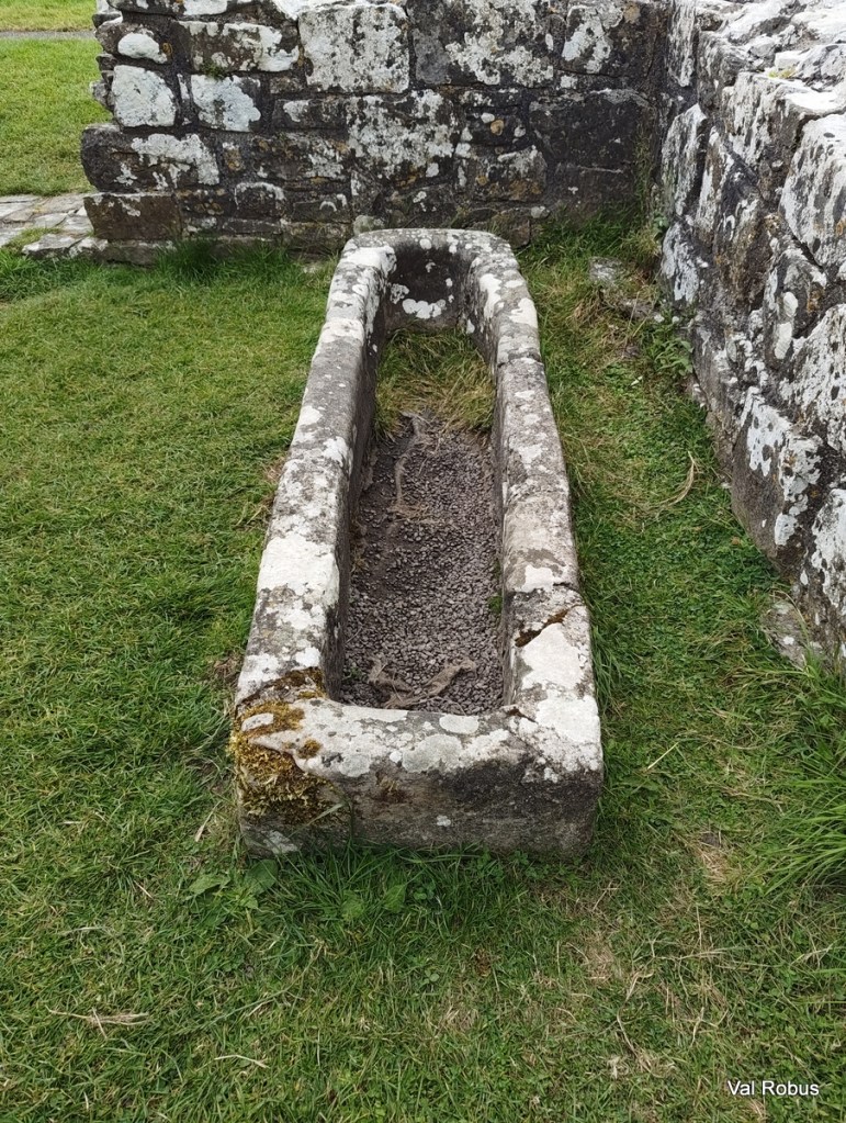 An old, stone trough surrounded by grass and a stone wall, showing signs of weathering and moss.