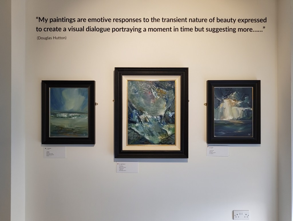 A gallery wall featuring three framed paintings by Douglas Hutton. The paintings depict abstract landscapes with varying colors and textures, alongside an inscription of Hutton's quote about his artistic approach.