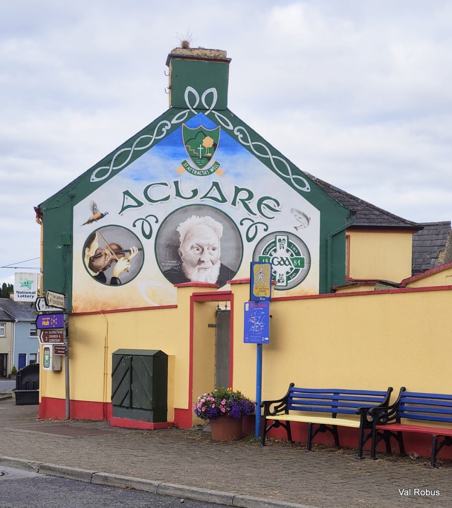 A colorful mural on a building in Aclare, featuring an elderly man's portrait, a violin, and local symbols. The wall is painted with green and yellow colors, and there are benches and flowerpots in front.