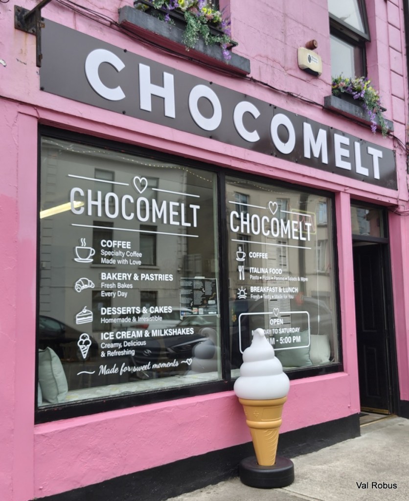 Exterior view of a cafe named 'Chocomelt,' featuring a pink facade and window signage showcasing menu items including coffee, pastries, desserts, and ice cream. A large decorative ice cream cone stands at the entrance.