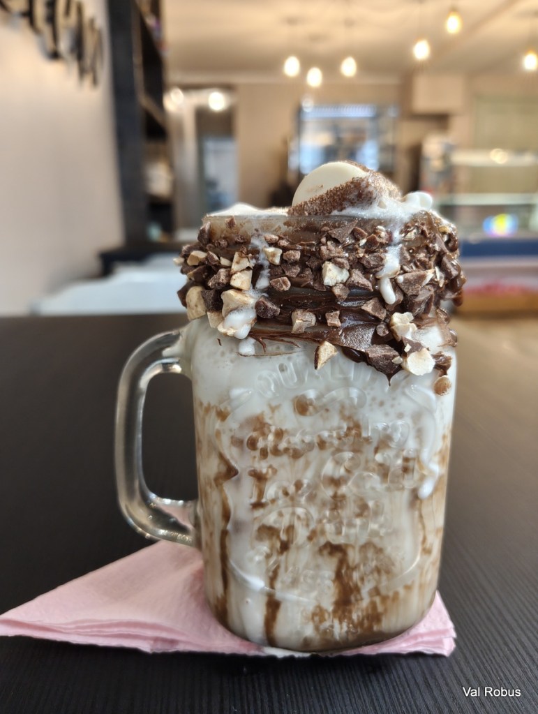 A glass mug filled with a chocolate milkshake topped with whipped cream, chocolate sauce, and chopped nuts, placed on a pink napkin against a blurred café background.