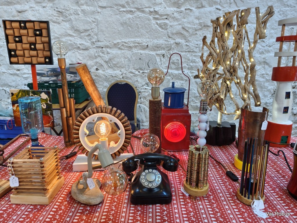A collection of unique, vintage-style lamps and home decor items displayed on a table covered with a patterned cloth, featuring a rotary phone, artistic light fixtures, and decorative elements made from wood, metal, and glass.