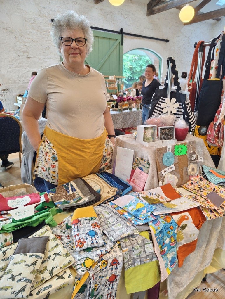 A smiling woman with curly grey hair and glasses stands behind a table filled with colorful handmade textiles, including aprons and household items, at a craft fair.