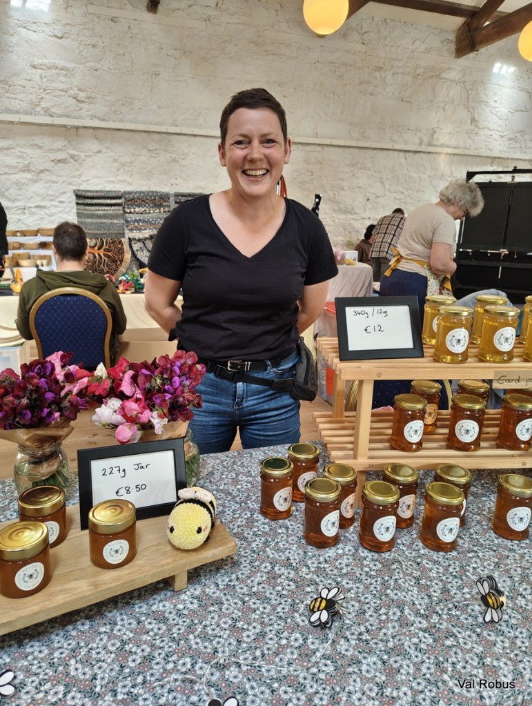 A smiling vendor stands behind a table displaying jars of honey and pricing signs at a market. The table also features decorative flowers and a crocheted bee toy.