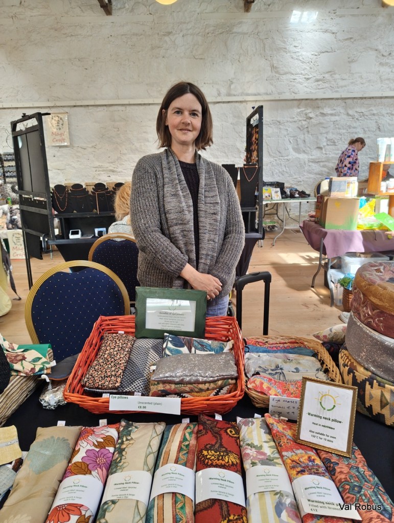 A woman stands behind a table displaying various handmade fabric items, including eye pillows and warming neck pillows, at a craft fair.
