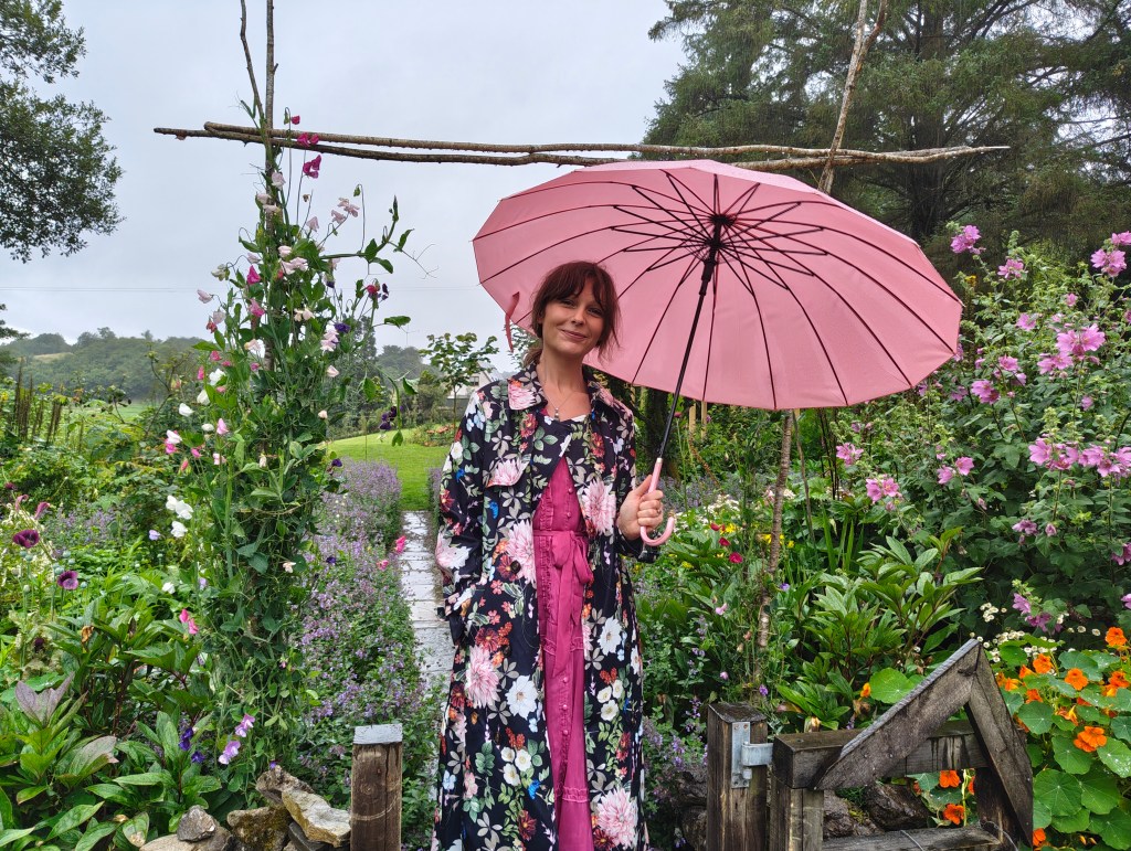 A woman standing in a vibrant garden filled with various flowers, holding a pink umbrella. She is wearing a floral coat over a pink dress, with greenery and colorful blooms surrounding her.