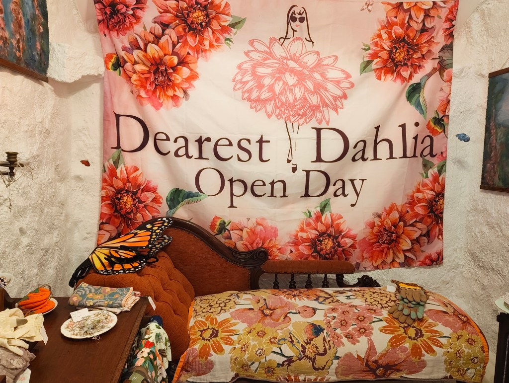 A cozy room featuring a large floral banner that reads 'Dearest Dahlia Open Day', with a vintage wooden bed adorned with a colorful floral quilt, decorative pillows, and various accessories on a bedside table.