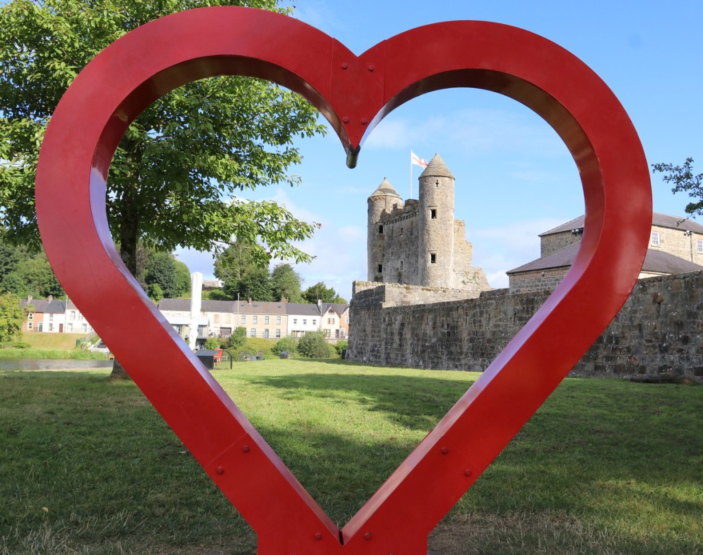 A large red heart sculpture in the foreground with a medieval castle in the background, surrounded by green grass and trees.