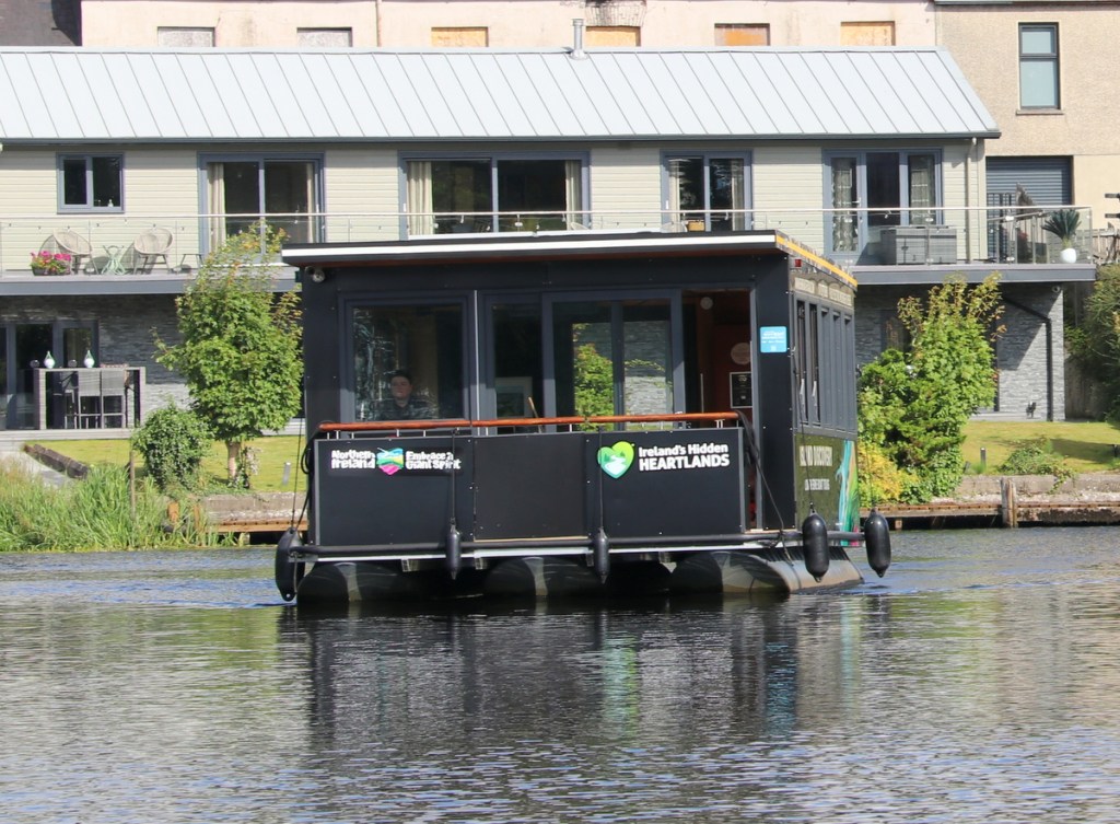 A black boat with a cabin cruising on a calm river, near modern homes and lush greenery.