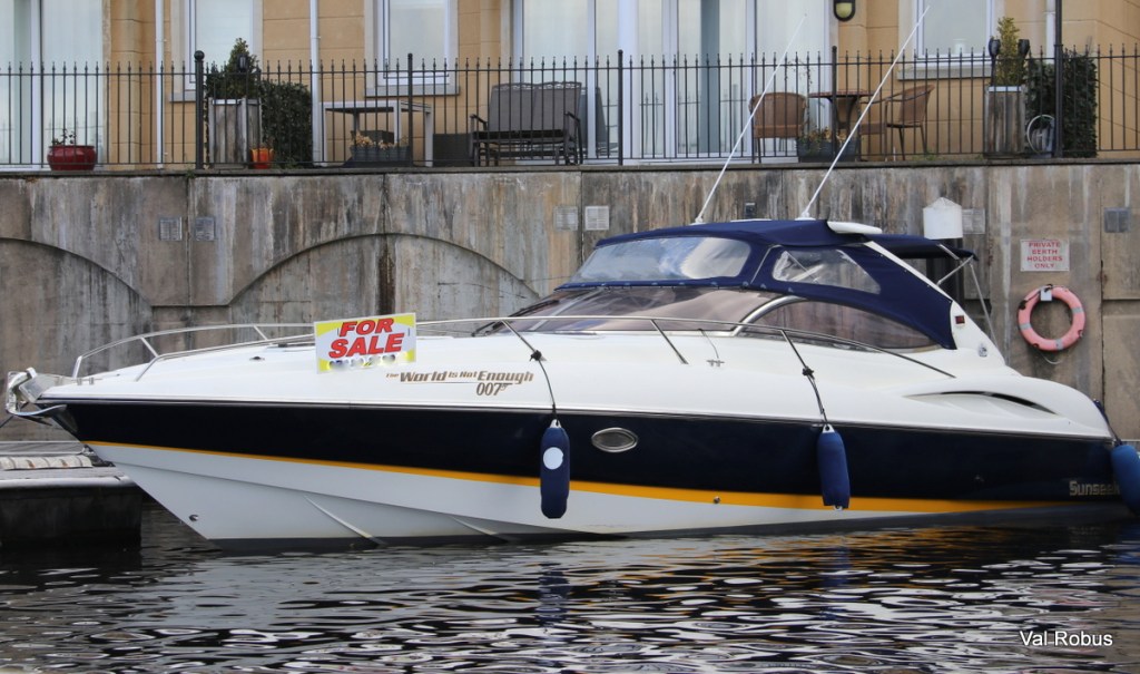 A motorboat with a blue hull and white upper deck, prominently displaying a 'FOR SALE' sign. The backdrop includes a stone wall and a railing, with a life ring visible nearby.