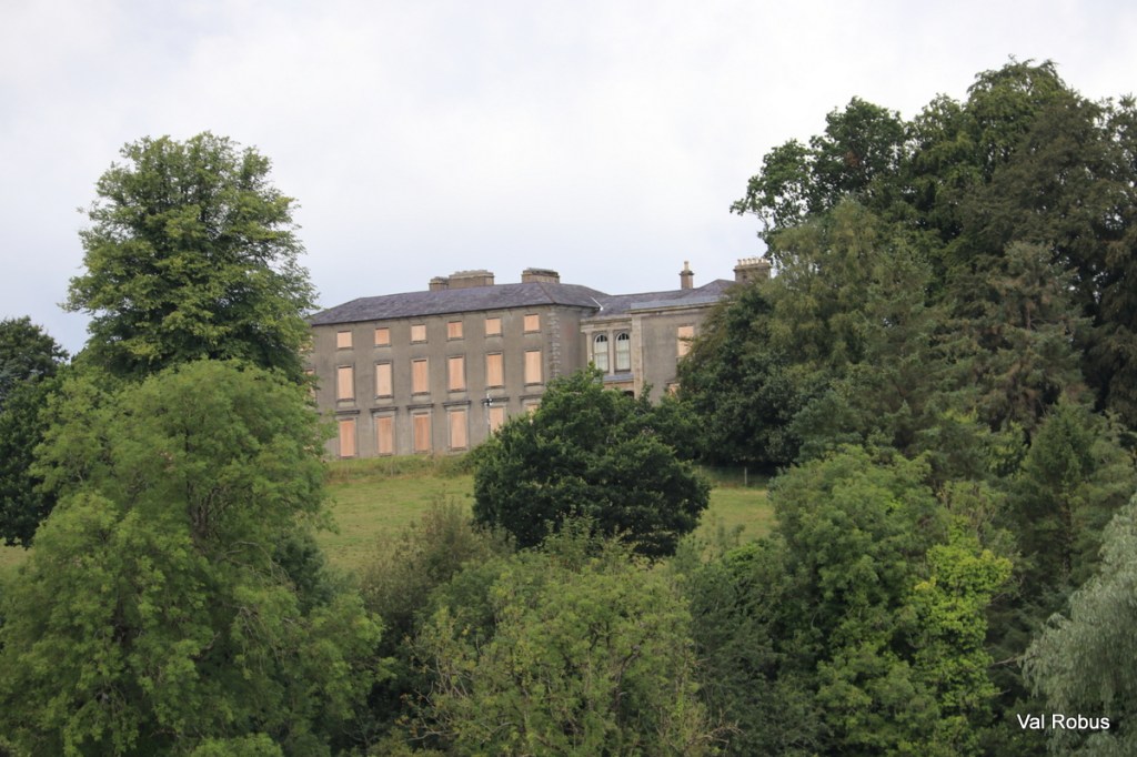 A historic building partially obscured by trees, situated on a hillside with cloudy skies in the background.
