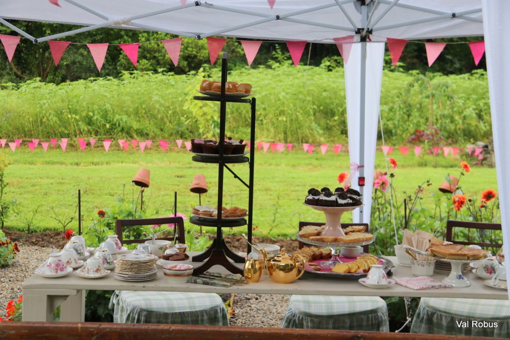 A charming outdoor setup featuring a dessert table under a white tent, adorned with pink bunting. The table displays an assortment of baked goods, including cupcakes, pastries, and cookies, alongside elegant tea cups and plates.