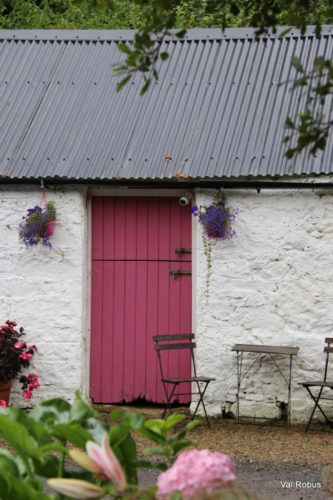 A charming white cottage with a pink door and a gray corrugated roof, featuring hanging flower pots and two metal chairs beside a small table.