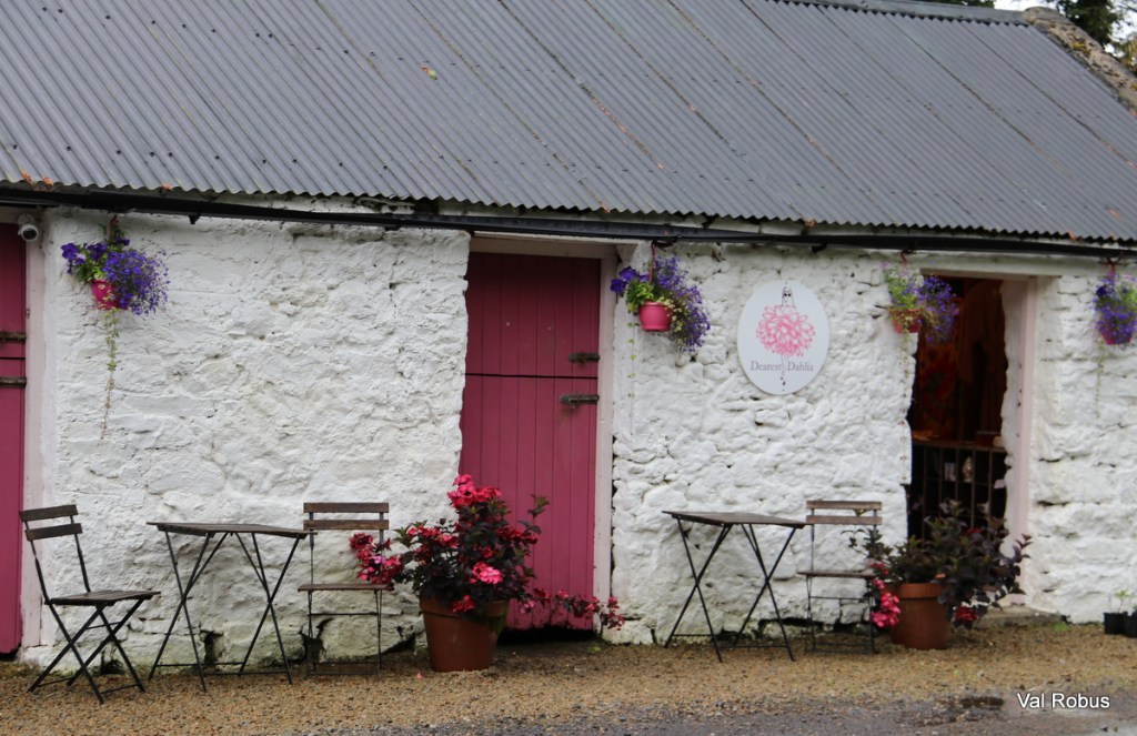 A charming white cottage with pink doors and a sign reading 'Daisy Dahlia.' The exterior features hanging flower pots and small tables with chairs arranged outside.